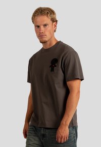 Camiseta de algodón gris con mangas cortas, que presenta una figura de dibujo animado negra bordada con un corazón, sobre un fondo neutro.