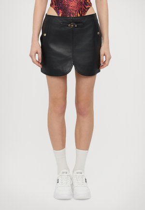 Jupe en cuir noir avec un ourlet courbé, comportant un bouton de fermeture à l'avant et des poches latérales. Portée avec des baskets blanches et des chaussettes.