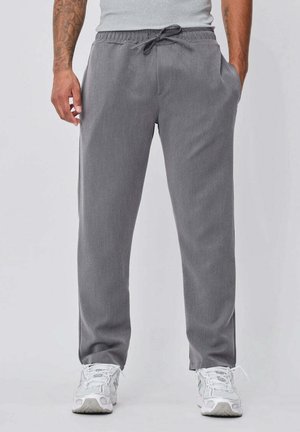 Homme portant un pantalon gris à cordon, des baskets blanches et un débardeur gris clair, debout les mains dans les poches.