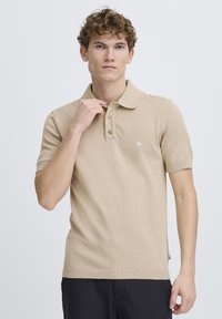 Polo din tricot bej cu mâneci scurte, plachetă cu trei nasturi și detaliu cu logo pe partea stângă a pieptului. Guler și tiv cu striații.