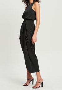 Vestido negro sin mangas con un diseño drapeado, que presenta una cintura ajustada y una falda fluida que cae por debajo de la rodilla, combinado con sandalias negras de tacón.