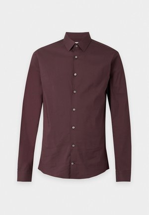 Ein Mann steht mit dem Gesicht nach vorne und trägt ein dunkel bordeauxfarbenes Hemd mit Knöpfen, hellbraune Hosen und schwarze Lederschuhe vor einem einfarbigen hellen Hintergrund.