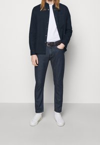 Tmavě modrá košile na knoflíky, vrstvená přes bílé tričko, doplněná o tmavé džíny a bílé tenisky. Akcentovaný opasek s jemným vzorem.