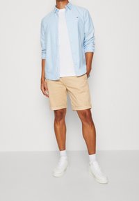 Ljusblå knapparskjorta över en vit t-shirt, matchat med beige shorts och vita sneakers. Avslappnad, figurnära design med uppvikta shorts.