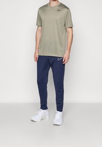 Olivově zelené tričko s krátkým rukávem Nike v kombinaci s tmavě modrými tepláky, které mají bílou Nike logo na levé noze, nošené s bílými teniskami.