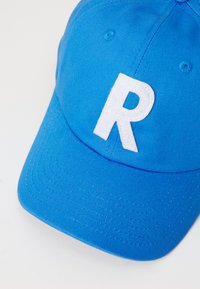 Blue baseball cap made of fabric featuring a white embroidered "R" on the front. Smooth texture with stitched detailing along the brim.