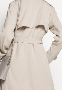 Beige trenchcoat med bälte knutet i midjan, en beige handväska med kedjestropp över axeln, visad bakifrån.