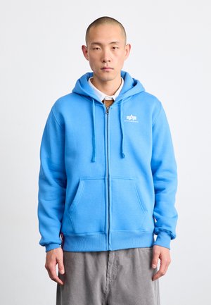 Felpa zip con cappuccio di colore azzurro chiaro, dotata di tasca frontale, con cordini e un logo ricamato sul lato sinistro del petto. Texture del tessuto morbida.