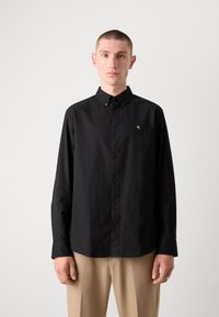 Black long-sleeve button-up shirt with a structured collar, featuring a subtle logo and textured fabric, paired with beige pants.