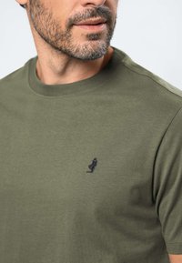 Homme à la barbe taillée portant un t-shirt vert olive avec une petite broderie noire de phare sur la poitrine, regardant sur le côté.
