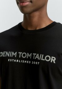 Musta T-paita, joka on valmistettu puuvillasta ja jossa on pyöreä pääntie sekä valkoisella painettu kuvio, jossa lukee "DENIM TOM TAILOR ESTABLISHED 2007."