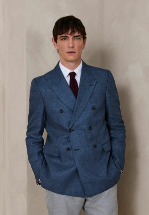 Mann steht mit den Händen in den Taschen, trägt einen blau karierten doppelt geknöpften Blazer, weißes Hemd, weinrote Krawatte und graue Hose.