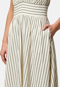 Kermakleid mit marineblauen vertikalen Streifen, strukturiertem Stoff, tailliertem Schnitt und Seitentasche. Faltener Rock sorgt für Bewegung und Fluss.