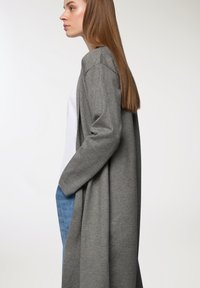 Długi szary kardigan z miękkiego materiału, z otwartym przodem, z opadającymi ramionami i luźnym krojem, noszony na białej bluzce i niebieskich dżinsach.