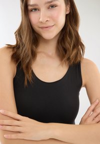 Giovane donna con pelle chiara e capelli castani ondulati, che indossa un top senza maniche nero, con le braccia incrociate, sorridendo dolcemente su uno sfondo semplice.