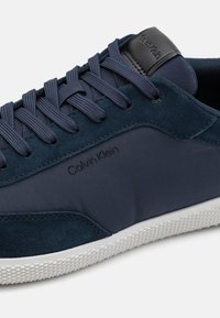 Marineblå sneakers laget av semsket skinn og stoff, med flate lisser, en logo på siden og en strukturert hvit gummisåle.