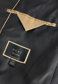 Black fabric with a hexagonal pattern, featuring a gold satin trim and a label that reads "NEXT Occasion Regular Fit 40R."
