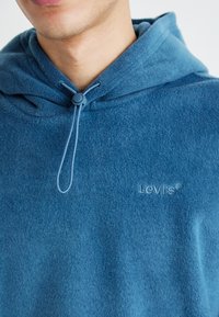 Felpa in pile blu con cappuccio regolabile, caratterizzata da un logo Levi's in rilievo sul petto. Texture morbida e design casual.