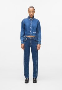 Korte denimjas met studs op de kraag en logo, gecombineerd met rechte jeans met studdetails langs de zijnaad. Donkerblauwe kleur.