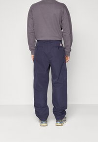 Homme portant un pantalon bleu marine ample, un sweat-shirt gris et des baskets beige, se tenant de dos devant un fond uni.