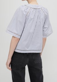 Personne portant un chemisier à rayures verticales bleues et blanches avec des manches courtes bouffantes et un jean noir taille haute, vue de dos.