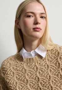 Beige gebreide trui met een textuurpatroon, voorzien van een ribbelneklijn. Een witte colbertlaag eronder toont dunne strepen.