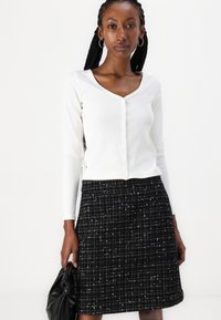 Haut long à manches blanches en maille côtelée avec un col en V et une fermeture à boutons, associé à une jupe en tweed noir et blanc présentant un motif à carreaux.