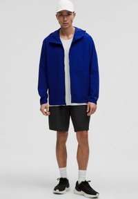 Man som bär en vit keps, blå uppdragbar jacka, vit skjorta, svarta shorts, vita strumpor och svarta sneakers, stående mot en enfärgad bakgrund.