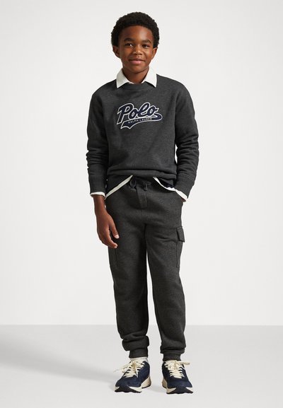 Pelēks sudraba krekls ar "Polo Ralph Lauren" logotipu, uzlikts virs baltas krekla ar apkakli; kombinācijā ar pelēkiem cargo treniņbiksēm un tumši ziliem sporta apaviem.