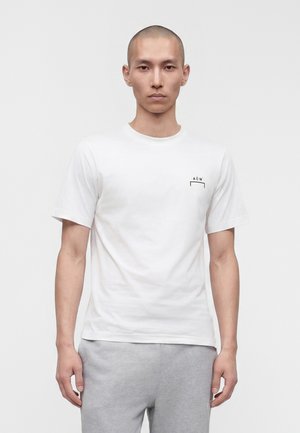 Man with shaved head wearing a white short-sleeve t-shirt with small black logo on chest and light gray pants, standing against white background.