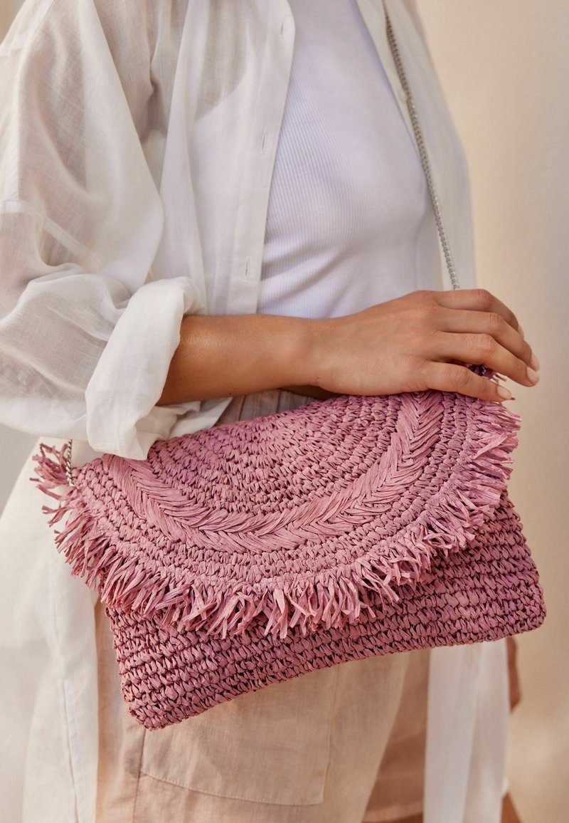 Femme portant une chemise blanche et un pantalon beige tenant une pochette tissée rose à franges avec une chaîne argentée.