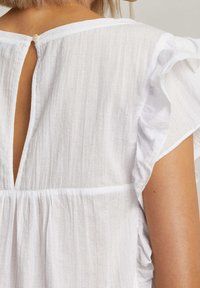 Vit texturerad blus med volangärmar, ett nyckelhål baktill med knappstängning, och en uppsamlad midja för en lös passform.