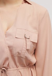 Ljusrosa blus i mjukt tyg, med V-urringning, en bröstficka med ögla och elastisk midja för passform.