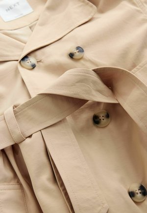 Beež trench mantel tekstuurse viimistlusega, uhkete suurte ümmarguste laiguliste nuppude ja sobiva vööga. Klassikalise krae kujundusega, volditud detailidega.