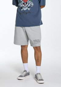 Pantaloni corti grigi chiari in felpa con un design grafico nero, abbinati a una maglietta grafica blu e sneakers grigie con dettagli a contrasto.