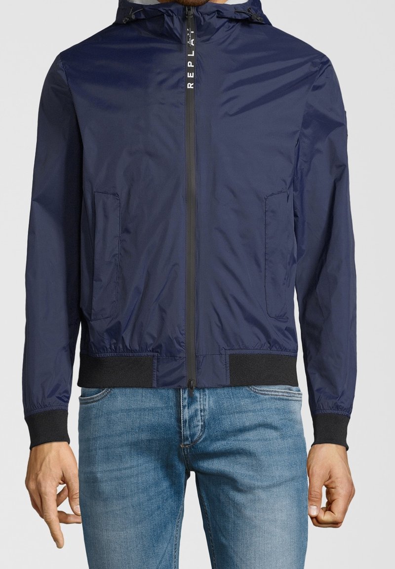 Giacca zip con cappuccio blu navy con polsini e vita neri, indossata con jeans azzurri, mostrata dalle spalle ai fianchi.