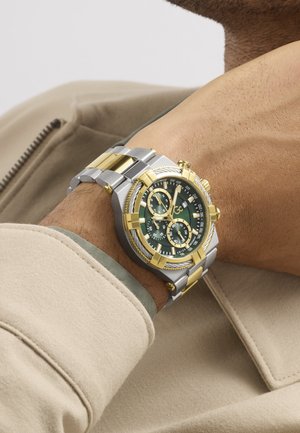 Náramkové hodinky so zlatým a strieborným kovovým remienkom a zeleným ciferníkom s viacerými subciferníkmi, nosené na ľavom zápästí osoby v béžovom saku.