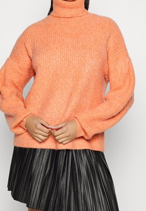 Frau trägt einen leuchtend orangefarbenen grob gestrickten Rollkragenpullover mit Ballonärmeln, kombiniert mit einem schwarzen Faltenrock.