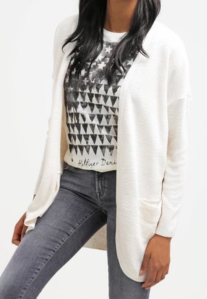 Femme portant un cardigan blanc côtelé ouvert sur un t-shirt graphique et un jean gris foncé, debout avec une main près de sa cuisse.