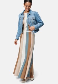 Pletené maxi šaty s vertikálnymi pruhmi v béžovej, hnedej a modrej farbe, doplnené svetlomodrou džínovou bundou. Biele lodičky zvýrazňujú outfit.