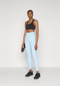 Must musta spordirinnahoidja, hele sinised kõrge vöökohaga retuusid ja mustad spordijalatsid. Modell hoiab veepudelit, juuksed on punutud. Siledad tekstuurid.