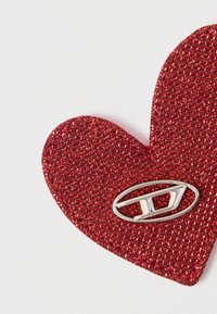 Cœur en tissu pailleté rouge avec une petite broche métallique argentée en forme d'ovale, arborant un design moderne en "D".