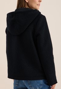 Veste en polaire noire avec capuche, en tissu texturé et coupe décontractée. Le dos présente un ourlet droit et des coutures minimales.