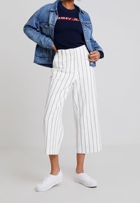 Persona che indossa pantaloni bianchi a vita alta con strisce verticali nere, sneakers bianche, maglione blu scuro e giacca di jeans blu, in piedi contro uno sfondo neutro.