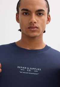 Joven con trenzas que lleva una camiseta azul marino con el texto "REPAIR & SUPPLIES NYC. CO. USA 'NUNCA DECEPCIONAMOS'" en el pecho.