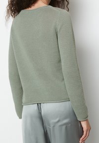 Personne portant un pull vert sauge texturé à manches longues et un pantalon en satin gris clair, vue de dos contre un mur blanc uni.