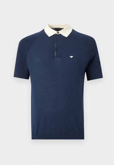 Κοντομάνικο μπλε ναυτικό πόλο με λευκό γιακά, τρία μαύρα κουμπιά και μικρό λευκό λογότυπο στο αριστερό στήθος.
