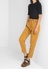 Svart ribbad kortärmad topp som kombineras med högmidjade senapsgula byxor med ett knytbälte, och svarta platsandaletter med randiga sulor.