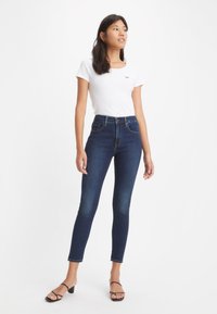 Schlank geschnittene dunkelblaue Jeans mit hoher Taille, kombiniert mit einem weißen T-Shirt mit einem kleinen Logo. Das Modell trägt schwarze Absatzsandalen.