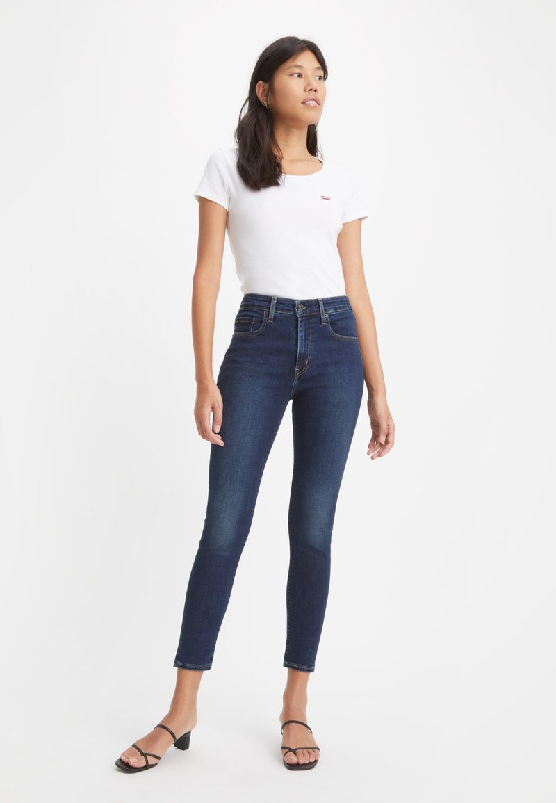Schlank geschnittene dunkelblaue Jeans mit hoher Taille, kombiniert mit einem weißen T-Shirt mit einem kleinen Logo. Das Modell trägt schwarze Absatzsandalen.